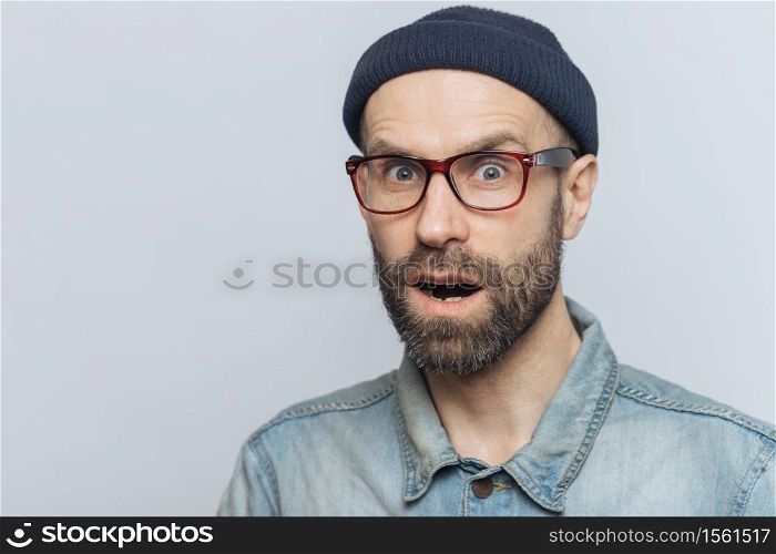 Suprised terrified unshaven male stares at camera and keeps mouth widely opened, wears fashionable clothing, expresses great shock as hears unexpected news from interlocutor, isolated on grey wall