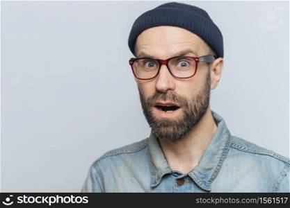 Suprised terrified unshaven male stares at camera and keeps mouth widely opened, wears fashionable clothing, expresses great shock as hears unexpected news from interlocutor, isolated on grey wall
