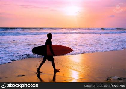 Surfer with surfboard walking on the sandy beach at sunset. Bali island, Indonesia