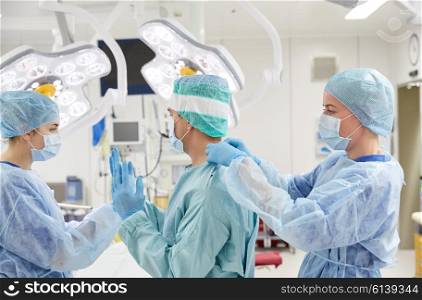 surgery, medicine and people concept - nurse assisting surgeon and helping with gloves and protective wear in operating room at hospital