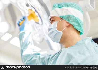 surgery, medicine and people concept - surgeon in mask adjusting lamp in operating room at hospital