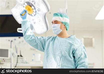surgery, medicine and people concept - surgeon in mask adjusting lamp in operating room at hospital