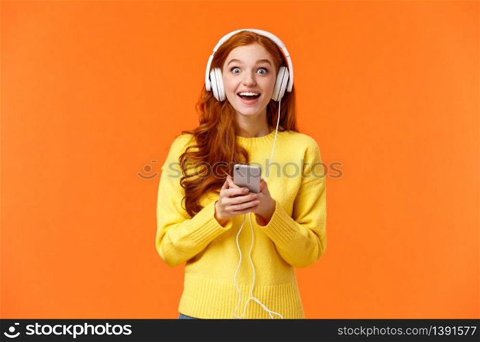 Surprised and astonished, excited smiling redhead girl found out favorite band released new song, listen music with fascinated, admiring eyes, grinning, holding smartphone, wear headphones.. Surprised and astonished, excited smiling redhead girl found out favorite band released new song, listen music with fascinated, admiring eyes, grinning, holding smartphone, wear headphones