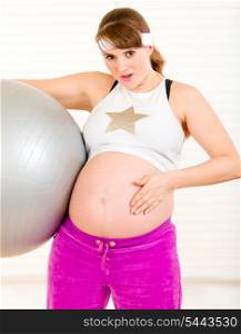 Surprised beautiful pregnant female holding fitness ball and touching her belly at living room&#xA;