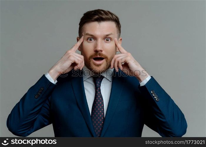 Surprised businessman in suit holding fingers on his temples while receiving shocking news, emotional male entrepreneur looking at camera with opened mouth while standing against grey background. Amazed businessman in suit holding fingers on temples and looking at camera with shocked face expression