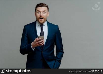Surprised businessman in suit holding smartphone and looking at camera with shocked face expression, male ceo executive reading unexpected news on mobile phone while standing against grey background. Surprised businessman in suit reading unexpected news on mobile phone