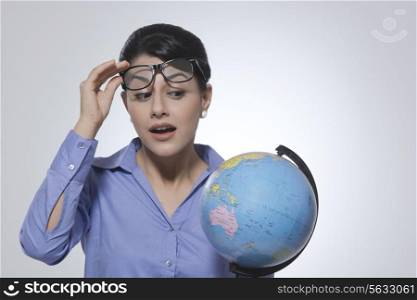 Surprised businesswoman looking at globe over glasses against gray background