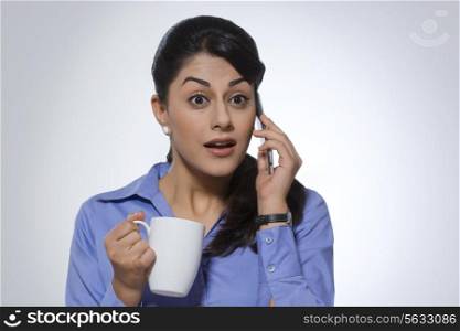 Surprised businesswoman with coffee mug using phone against gray background
