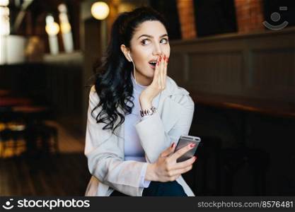 Surprised elegant lady with dark hair having long red nails holding hand on her full lips looking aside using smartphone and earphones resting at cafe. Cute female using modern gadget indoors