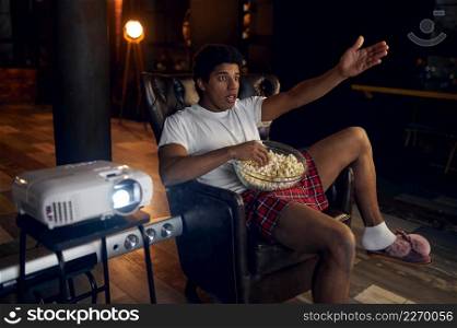 Surprised emotional man with mouth full of popcorn watching film or show program on projector. Surprised indignant man watching film on projector