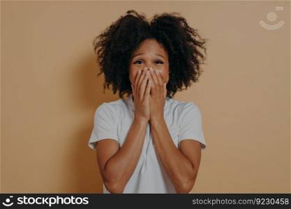 Surprised excited dark skinned woman in white tshirt covering her mouth with hands isolated on pastel beige background with copy space. Positive women emotions and body language concept. Surprised excited dark skinned woman in white tshirt covering her mouth with hands