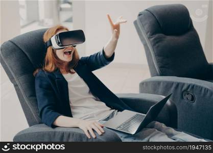 Surprised excited red haired woman in VR headset goggles and laptop, sitting in soft big armchair in office, trying to touch 3D virtual reality objects, pointing with index finger up in air. Surprised excited woman in VR headset goggles and laptop, sitting in armchair in office