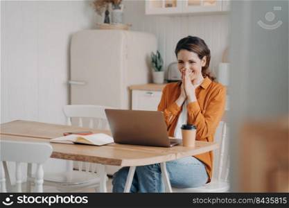 Surprised excited young woman is sitting in front of laptop having video call. Caucasian girl in orange shirt is remote worker at the kitchen at home. Distance work on quarantine concept.. Surprised excited young woman is sitting in front of laptop having video call. Distance work.