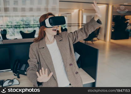 Surprised female office worker wearing VR headset surprised with how realistic virtual reality is, cannot contain emotions while testing 3d goggles at work, holding hands in front of her and gesturing. Woman wearing VR headset surprised with virtual reality while standing near her workplace in office