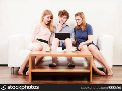 Surprised friends browsing surfing internet on tablet. Young people man guy and women girls sitting on sofa at home.. Friends relaxing browsing internet on tablet.