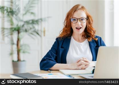 Surprised ginger female employee has job, develops new business srategy, poses in front of opened laptop computer, poses in workspace against office interior. Woman software developer at work.