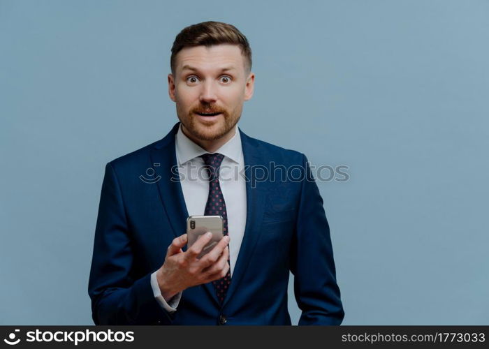 Surprised happy businessman or male entrepreneur in suit holding cell phone and getting good positive news, looking at camera with shocked face expression while standing against grey background. Surprised businessman reading good news on mobile phone