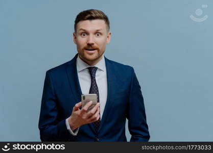 Surprised happy businessman or male entrepreneur in suit holding cell phone and getting good positive news, looking at camera with shocked face expression while standing against grey background. Surprised businessman reading good news on mobile phone