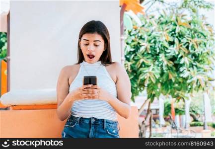 Surprised person looking at cell phone outdoor. Girl with cell phone with a surprised face leaning on a wall. Young woman with the cell phone with a gesture of surprise with open mouth outside
