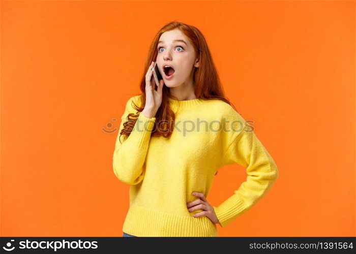 Surprised, startled good-looking redhead female hear amazing news as talking on phone, drop jaw stare left and holding smartphone near ear during conversation, standing orange background.. Surprised, startled good-looking redhead female hear amazing news as talking on phone, drop jaw stare left and holding smartphone near ear during conversation, standing orange background