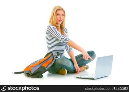 Surprised teenager sitting on floor with backpack and pointing in laptop isolated on white &#xA;