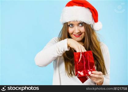 Surprised woman wearing santa claus hat puts her hand into christmas red gift bag, on blue. Christmas woman puts her hand into gift bag