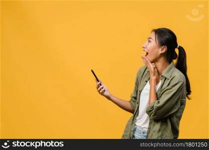 Surprised young Asia lady using mobile phone with positive expression, smiles broadly, dressed in casual clothing and stand isolated on yellow background. Happy adorable glad woman rejoices success.