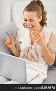 Surprised young housewife sitting on couch with laptop and credit card