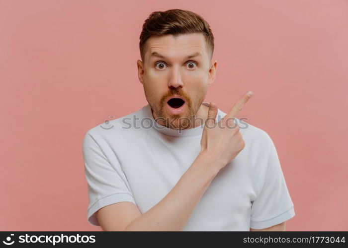 Surprised young unshaven guy in white shirt pointing with finger to side at copy space with amazed wow face expression, advertising something fantastic while standing isolated over pink background. Shocked young man pointing with finger to side with surprised face expression