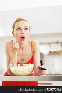 Surprised young woman eating popcorn and watching tv in kitchen