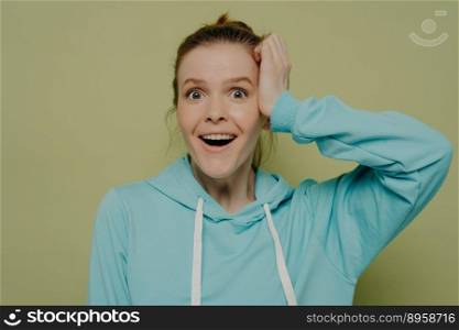 Surprised young woman without makeup demonstrating shock and excitement holding hand at head looking at camera wearing casual blue hooded sweater while posing in studio. Human emotions concept. Excited young woman expressing shock holding hand at head