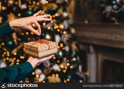 Surprisment and pleasant moments concept. Woman wrapps New Year gift as stands in living room near beautiful decorated fir tree. Holidays, x mas and celebration concept.