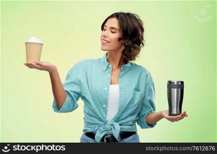 sustainability and eco living concept - portrait of smiling young woman comparing thermo cup or tumbler with disposable paper coffee cup over green background. woman comparing thermo cup and paper coffee cup
