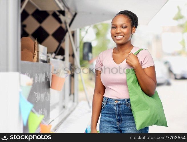 sustainability and people concept - happy smiling young african american woman with green reusable canvas bag for shopping over food truck background. woman with reusable bag over food truck