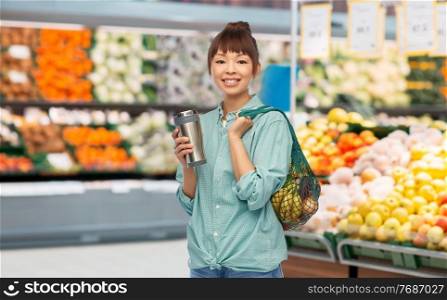 sustainability and people concept - portrait of happy asian woman in turquoise shirt with thermo cup or tumbler for hot drinks over grocery store background. woman with tumbler and food in bag at store