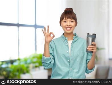 sustainability and people concept - portrait of smiling asian woman with thermo cup for hot drinks showing ok hand sign over home room background. woman with thermo cup or tumbler showing ok sign