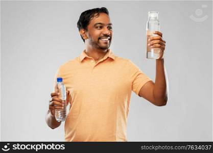 sustainability, consumerism and eco friendly concept - happy smiling indian man comparing water in reusable glass and plastic bottles grey background. indian man comparing water in different bottles