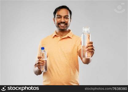 sustainability, consumerism and eco friendly concept - happy smiling indian man comparing water in reusable glass and plastic bottles grey background. indian man comparing water in different bottles