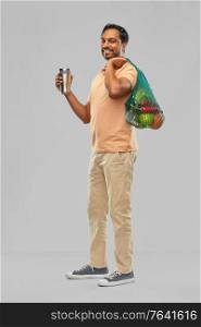 sustainability, consumerism and eco friendly concept - happy smiling indian man holding green reusable string bag for food shopping and tumbler or thermo cup on grey background. man with food in bag and tumbler or thermo cup