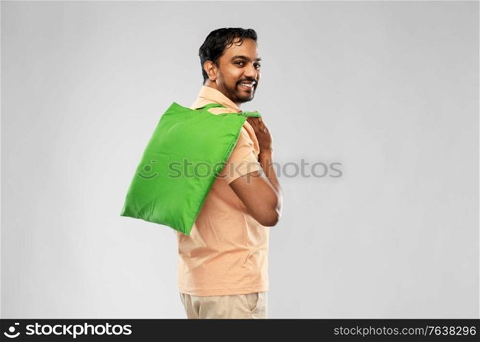 sustainability, consumerism and eco friendly concept - happy smiling indian man with green reusable canvas bag for food shopping on grey background. man with reusable canvas bag for food shopping