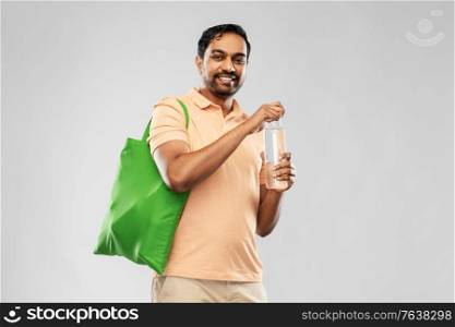 sustainability, consumerism and eco friendly concept - happy smiling indian man with green reusable canvas bag for food shopping and glass bottle of water on grey background. man with bag for food shopping and glass bottle
