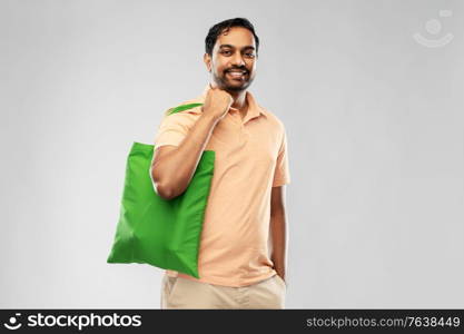 sustainability, consumerism and eco friendly concept - happy smiling indian man with green reusable canvas bag for food shopping on grey background. man with reusable canvas bag for food shopping