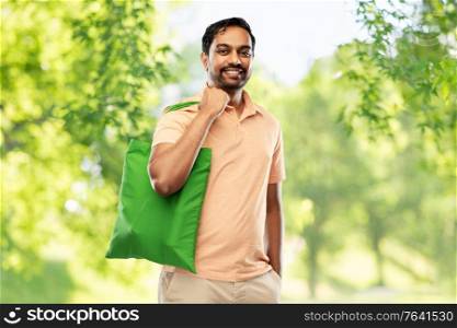 sustainability, consumerism and eco friendly concept - happy smiling indian man with green reusable canvas bag for food shopping over natural background. man with reusable canvas bag for food shopping