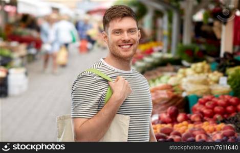 sustainability, consumerism and eco friendly concept - happy smiling man with reusable canvas bag for food shopping over street market on background. man with reusable shopping bag at street market