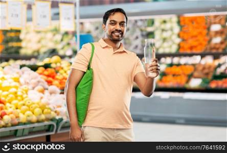 sustainability, consumerism and eco friendly concept - smiling indian man with green reusable canvas bag for food shopping and glass bottle of water over supermarket or grocery store on background. man with bag for food shopping and glass bottle