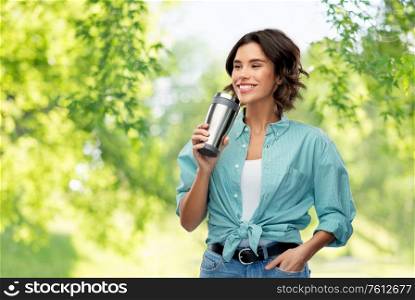 sustainability, eco and green living concept - portrait of happy smiling young woman in turquoise shirt drinking from thermo cup or tumbler for hot drinks over natural background. woman drinking from thermo cup or tumbler