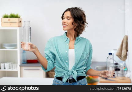 sustainability, eco and green living concept - portrait of happy smiling young woman in turquoise shirt comparing water in plastic and reusable glass bottle grey background. smiling young woman comparing bottles of water