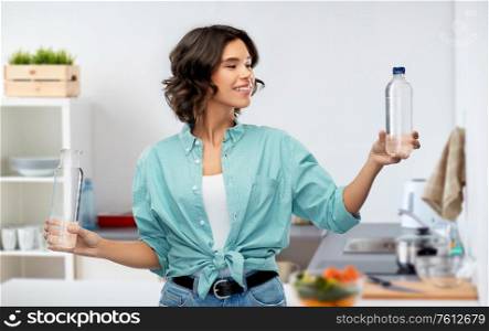 sustainability, eco and green living concept - portrait of happy smiling young woman in turquoise shirt comparing water in plastic and reusable glass bottle grey background. smiling young woman comparing bottles of water