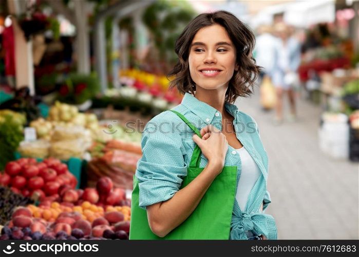 sustainability, eco and green living concept - portrait of happy smiling young woman in turquoise shirt with reusable canvas bag for food shopping over street market background. woman with reusable canvas bag for food shopping