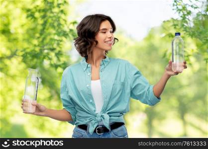 sustainability, eco and green living concept - portrait of happy smiling young woman in turquoise shirt comparing water in plastic and reusable glass bottle over green natural background. smiling young woman comparing bottles of water